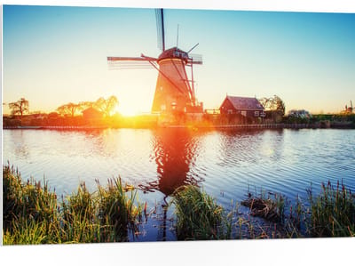 WallClassics - PVC Schuimplaat- Zonlicht langs Hollandse Molen aan het Water - 105x70 cm Foto op PVC Schuimplaat